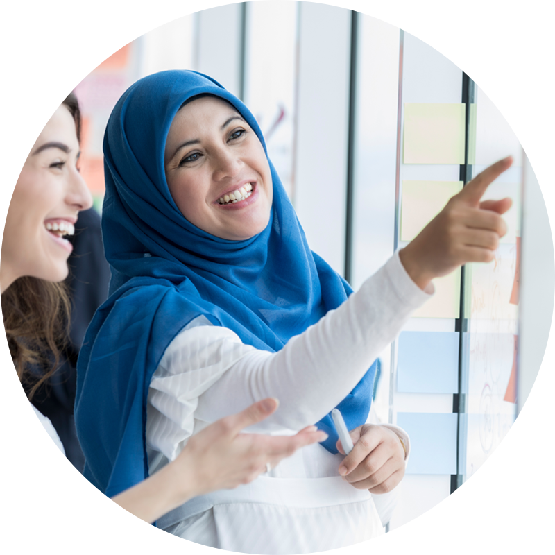 Une femme d'affaires d'âge moyen, heureuse et portant un hijab, montre du doigt une idée qu'elle a notée sur un post-it. Son associée sourit en discutant de l'idée.